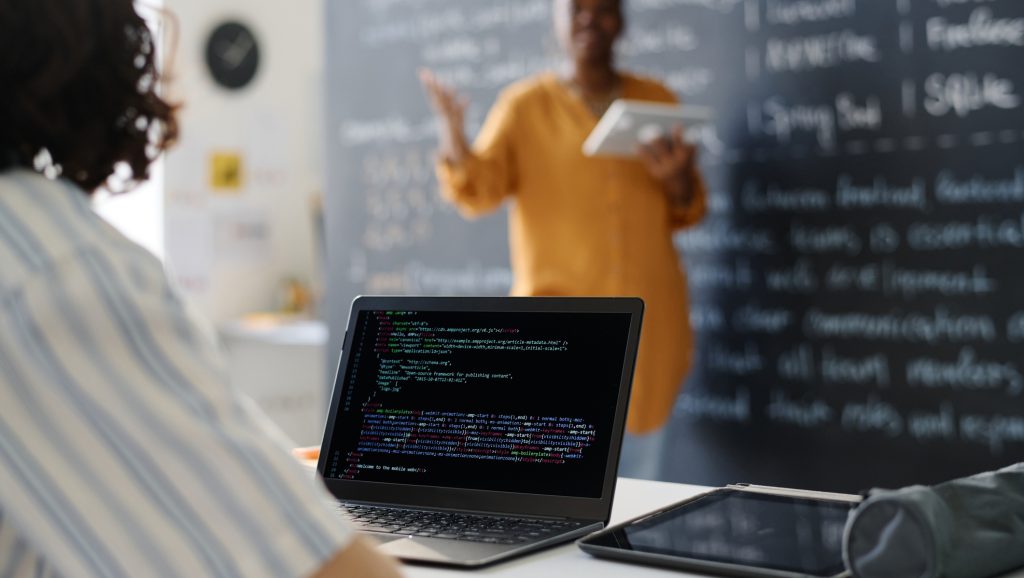 Rear view of student typing codes on computer while sitting at desk at lesson with teacher explaining material in background